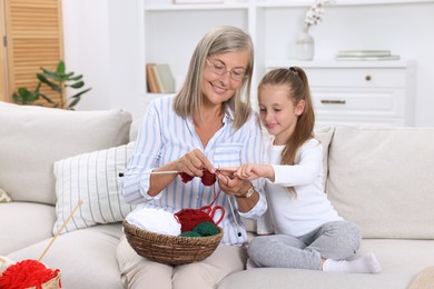 Photo of Smiling grandmother teaching her granddaughter to knit on sofa at home