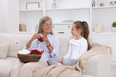 Photo of Smiling grandmother teaching her granddaughter to knit on sofa at home