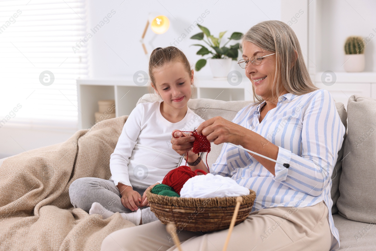 Photo of Smiling grandmother teaching her granddaughter to knit on sofa at home