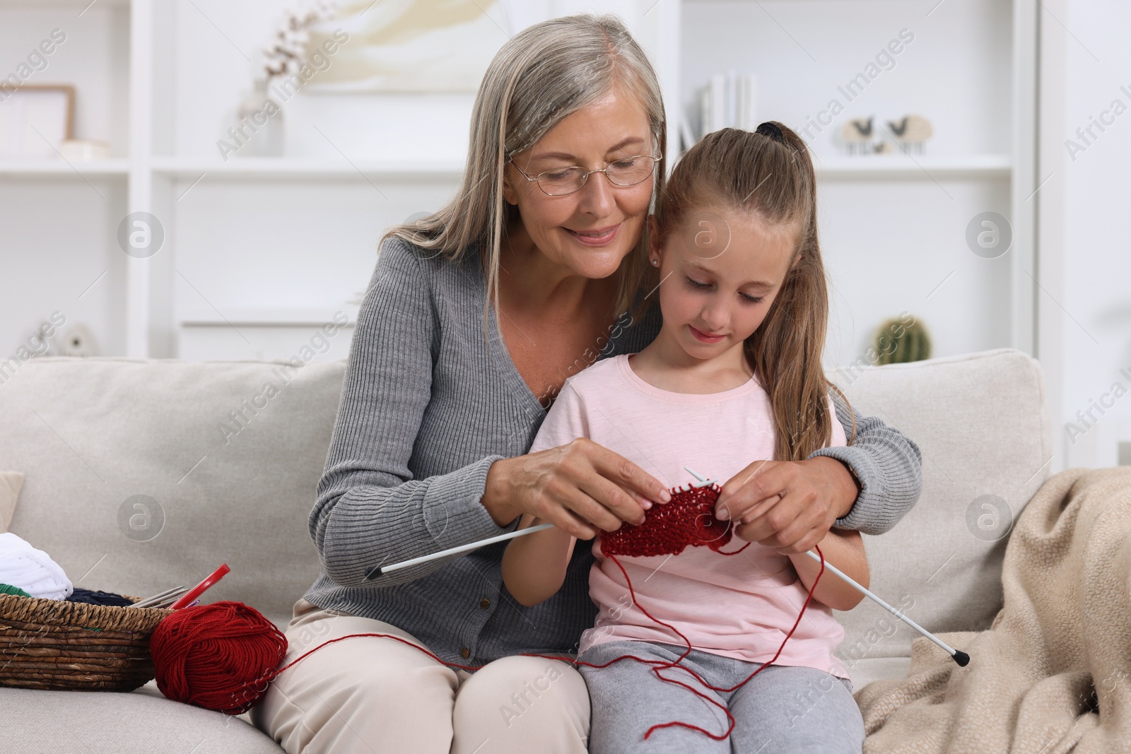 Photo of Smiling grandmother teaching her granddaughter to knit on sofa at home