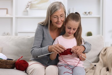 Smiling grandmother teaching her granddaughter to knit on sofa at home