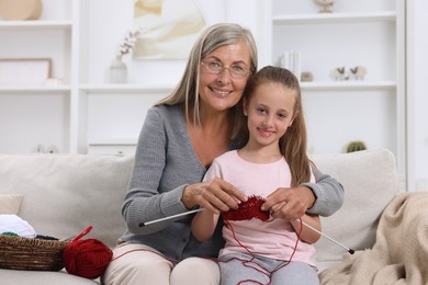 Smiling grandmother teaching her granddaughter to knit on sofa at home