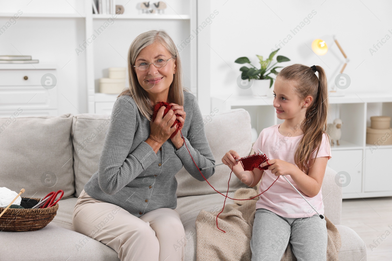 Photo of Grandmother teaching her granddaughter to knit on sofa at home