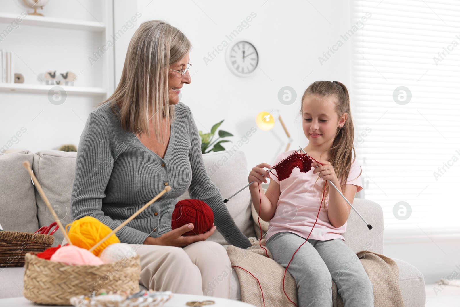 Photo of Smiling grandmother teaching her granddaughter to knit on sofa at home