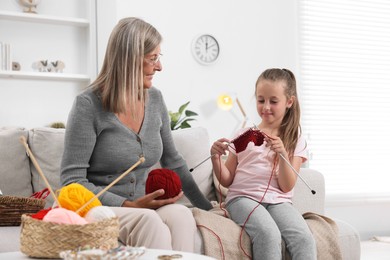 Photo of Smiling grandmother teaching her granddaughter to knit on sofa at home