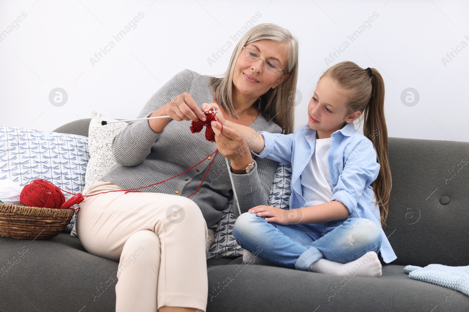 Photo of Grandmother teaching her granddaughter to knit on sofa at home