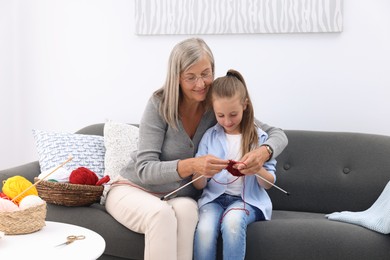 Photo of Smiling grandmother teaching her granddaughter to knit on sofa at home