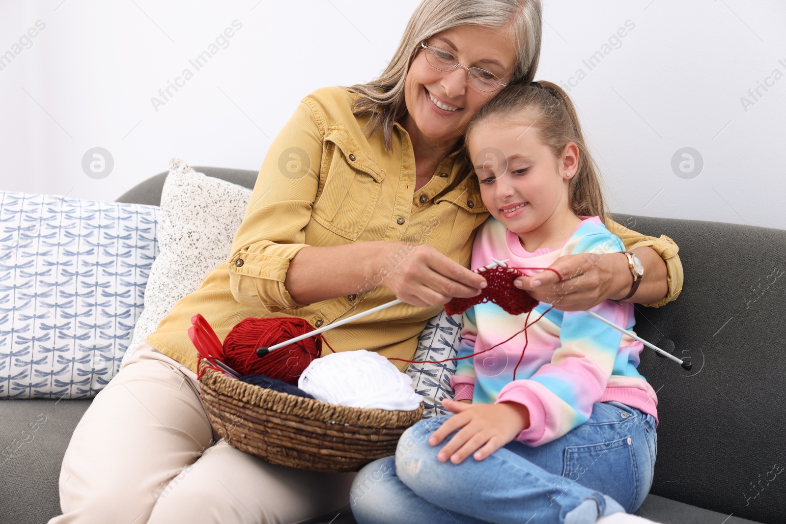 Photo of Smiling grandmother teaching her granddaughter to knit on sofa at home