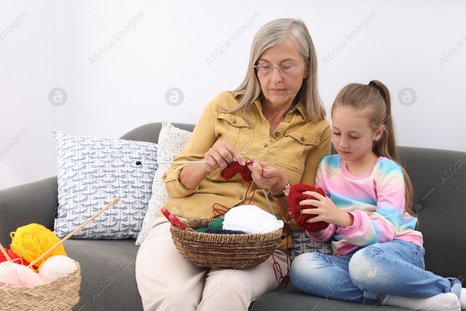 Photo of Grandmother teaching her granddaughter to knit on sofa at home