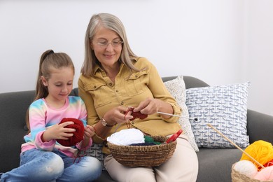 Photo of Grandmother teaching her granddaughter to knit on sofa at home