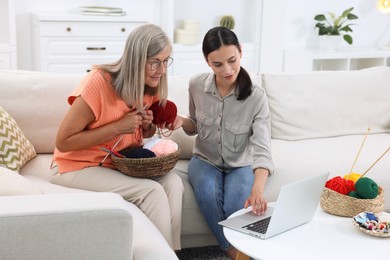 Mother and daughter watching video tutorial via laptop how to knit on sofa at home