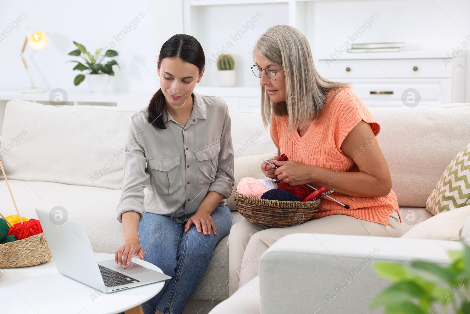 Photo of Mother and daughter watching video tutorial via laptop how to knit on sofa at home