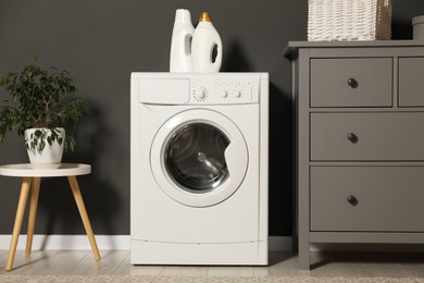 Photo of Washing machine, detergents, chest of drawers and houseplant on table indoors