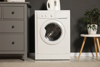 Photo of Washing machine, detergents, chest of drawers and houseplant on table indoors