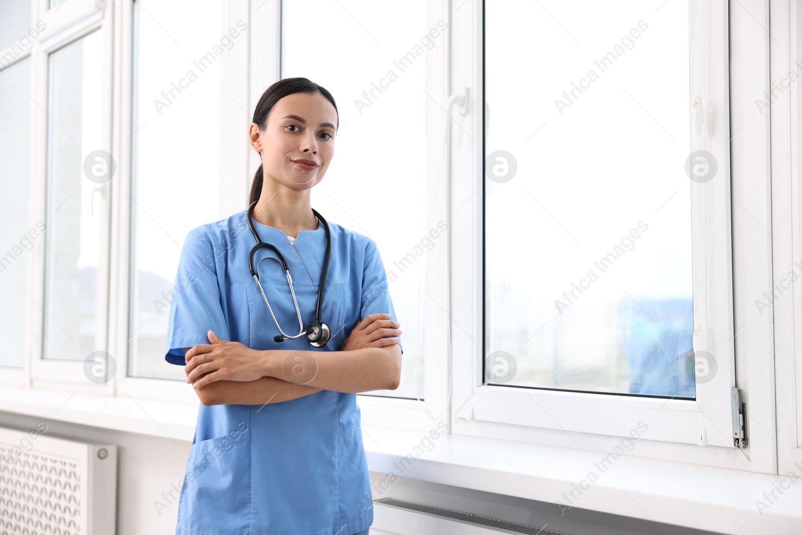 Photo of Beautiful nurse with crossed arms near window in clinic. Space for text