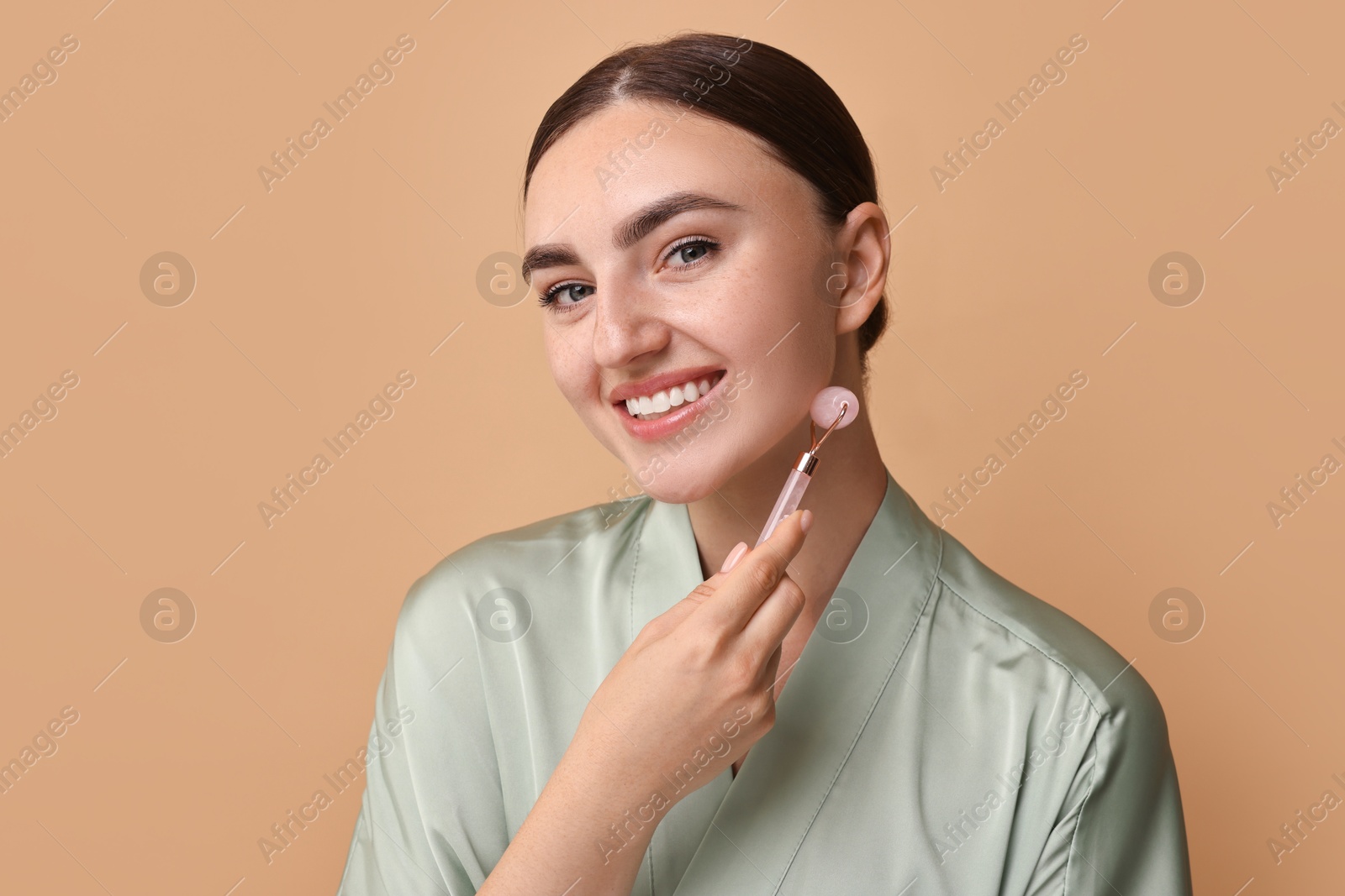 Photo of Beautiful young woman doing facial massage with roller on beige background