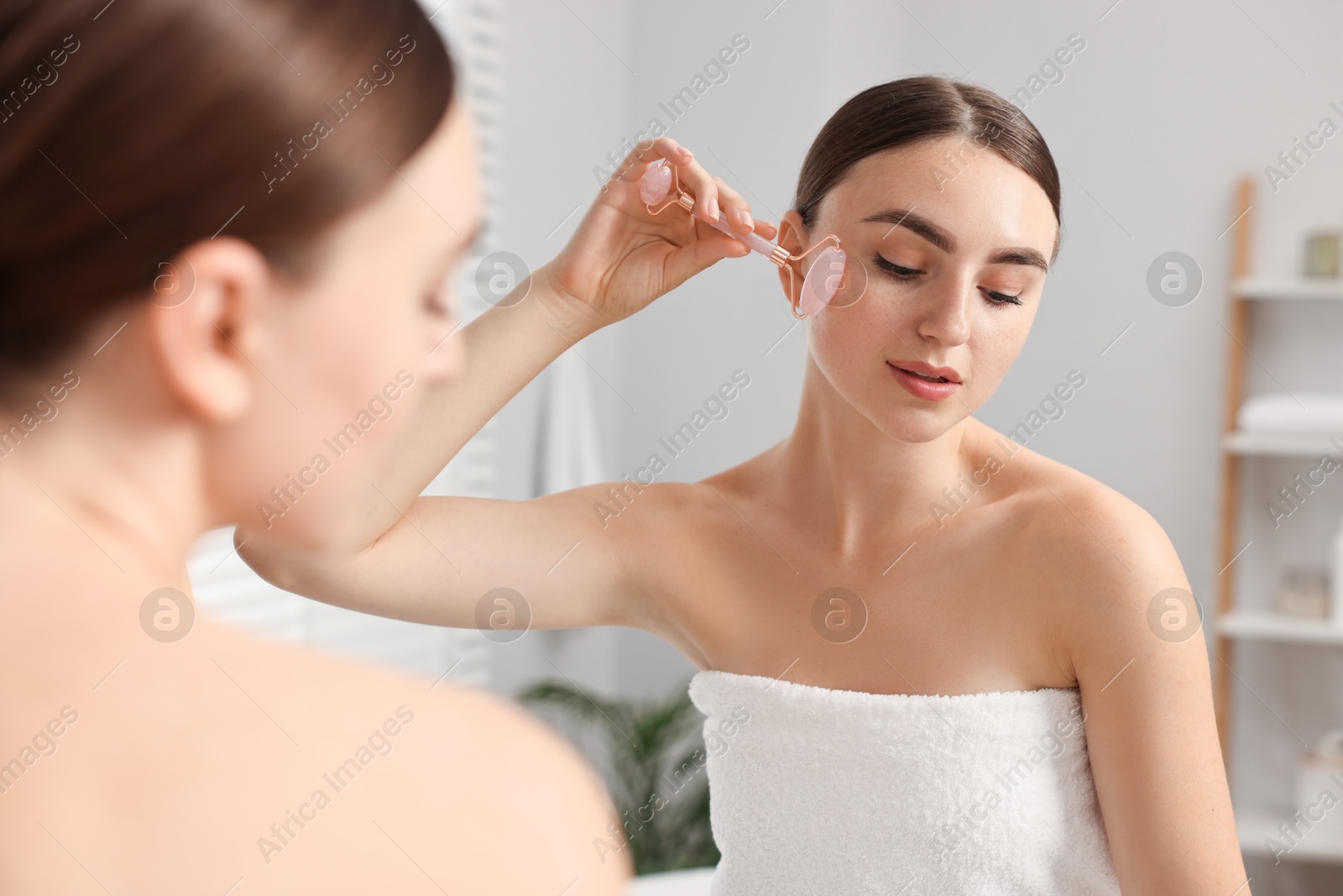 Photo of Beautiful young woman doing facial massage with roller near mirror at home