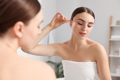 Photo of Beautiful young woman doing facial massage with roller near mirror at home