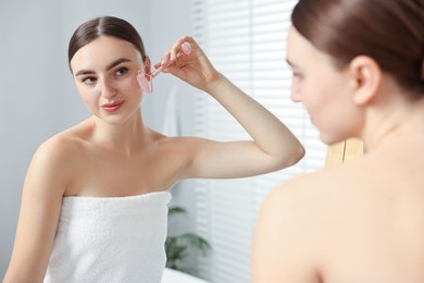 Photo of Beautiful young woman doing facial massage with roller near mirror at home