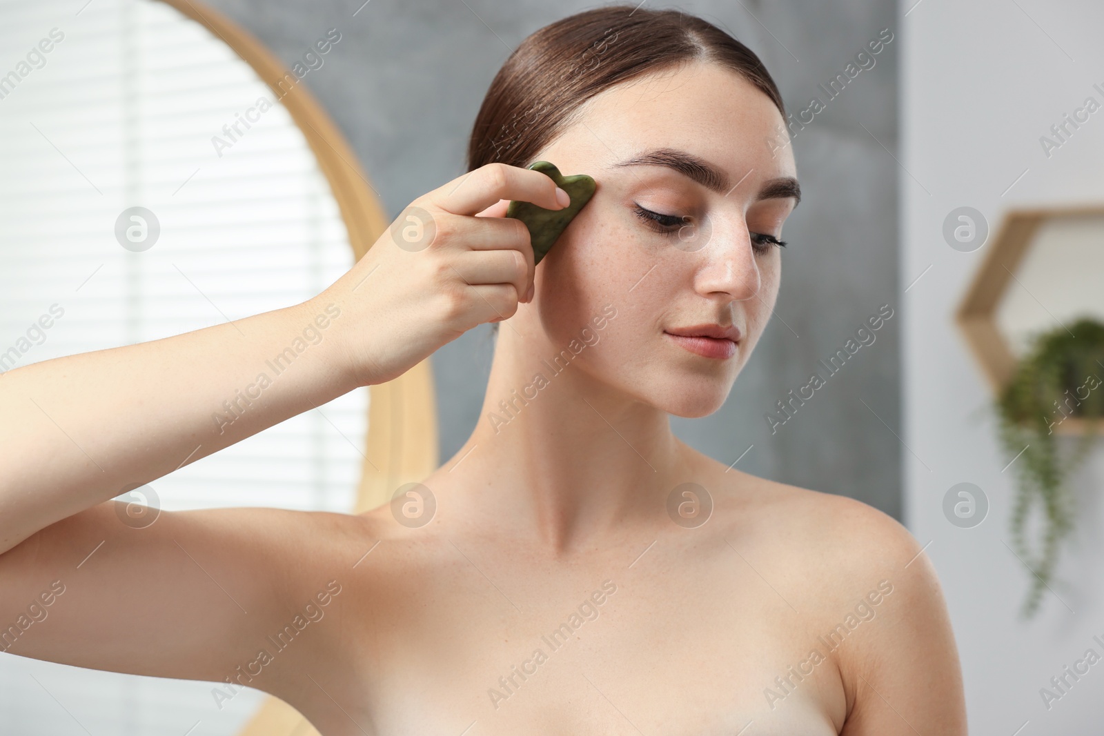 Photo of Beautiful young woman doing facial massage with gua sha tool indoors