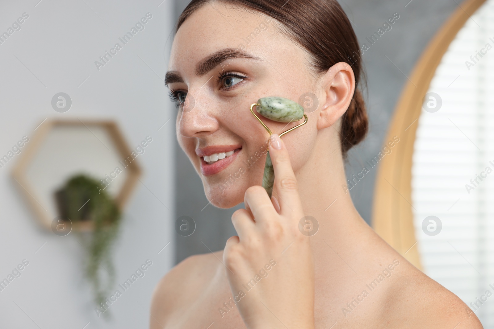 Photo of Beautiful young woman doing facial massage with roller indoors