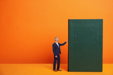 Small businessman touching huge book on orange background, space for text. Concept of education and knowledge