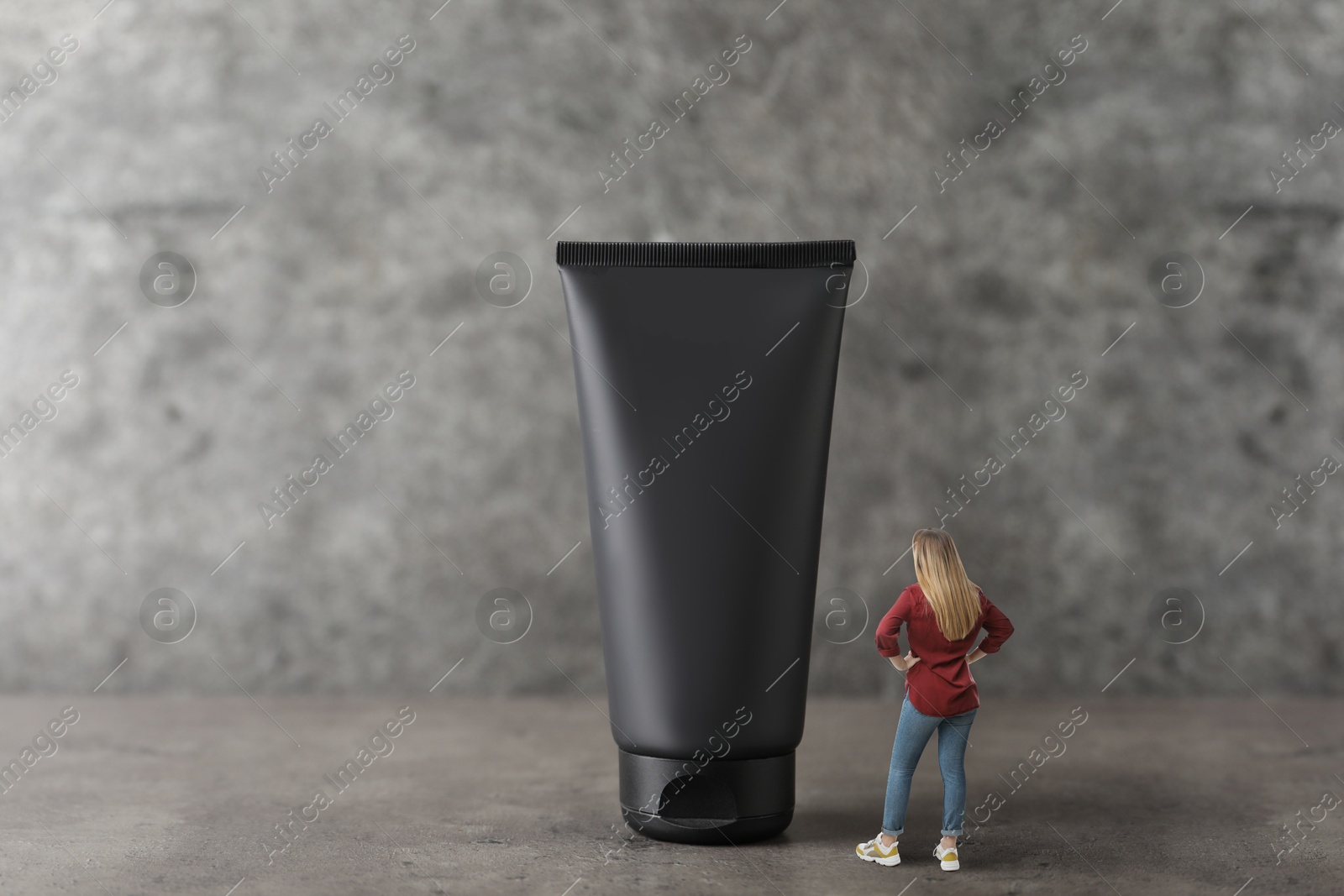 Image of Small woman looking at big black tube of cream on grey background. Concept of beauty care