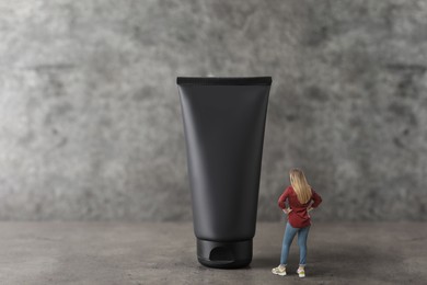 Image of Small woman looking at big black tube of cream on grey background. Concept of beauty care