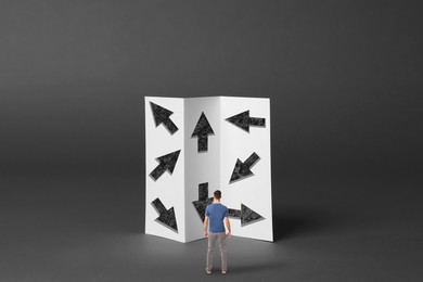 Small man looking at paper with arrows pointing in different directions on grey background. Concept of choice