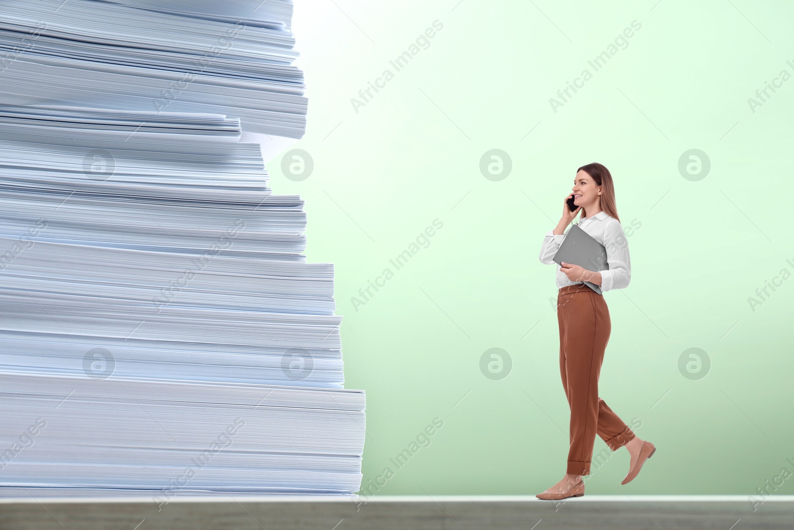 Image of Confident businesswoman talking on phone while walking towards huge stack of papers against light green background. Concepts of success, ambition