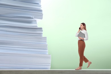 Image of Confident businesswoman talking on phone while walking towards huge stack of papers against light green background. Concepts of success, ambition