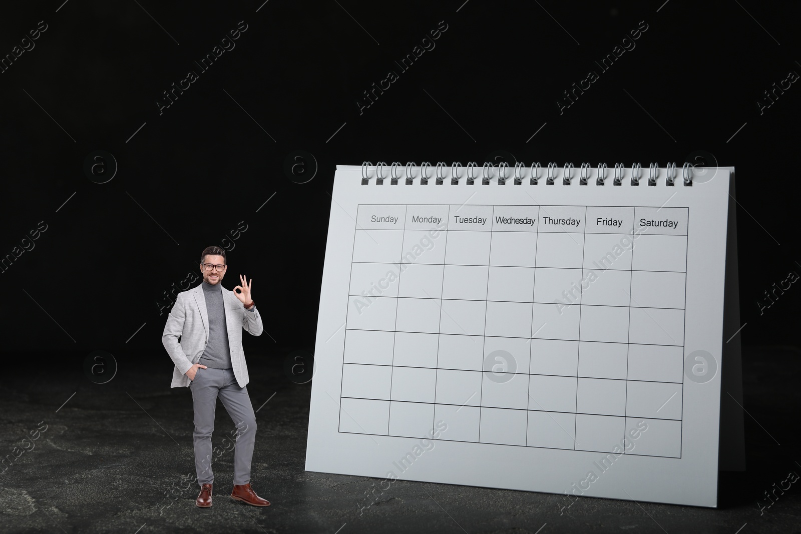 Image of Happy small businessman showing Ok sign near big calendar on black background. Concepts of self-management, planning