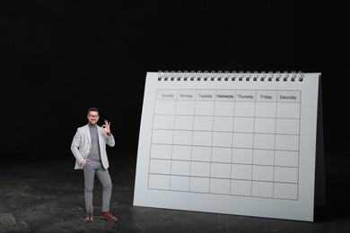 Image of Happy small businessman showing Ok sign near big calendar on black background. Concepts of self-management, planning