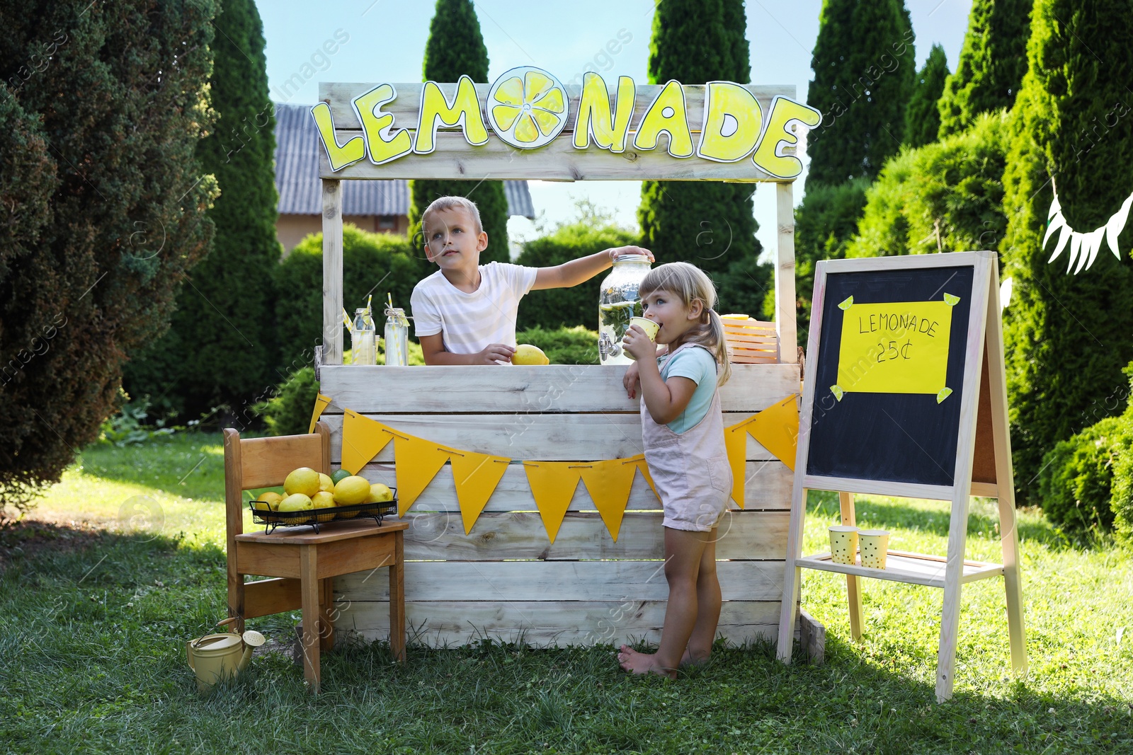 Photo of Cute boy standing at wooden stand while little girl drinking refreshing lemonade in park