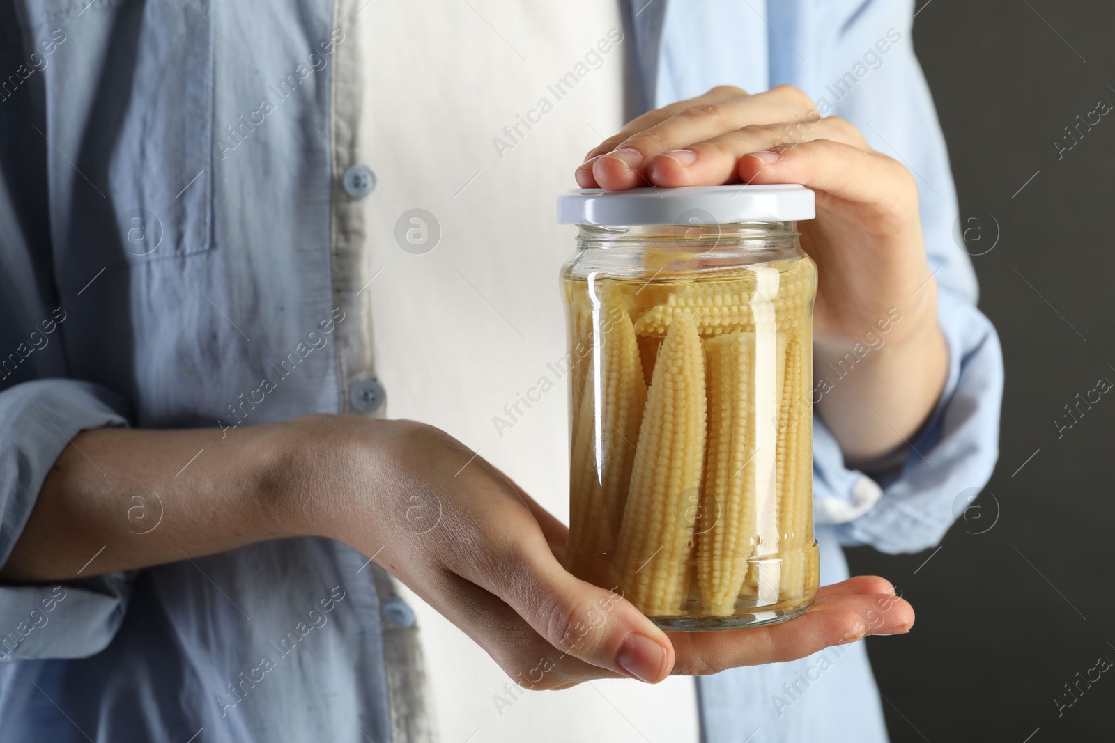 Photo of Woman with jar of tasty pickled yellow baby corns on gray background, closeup