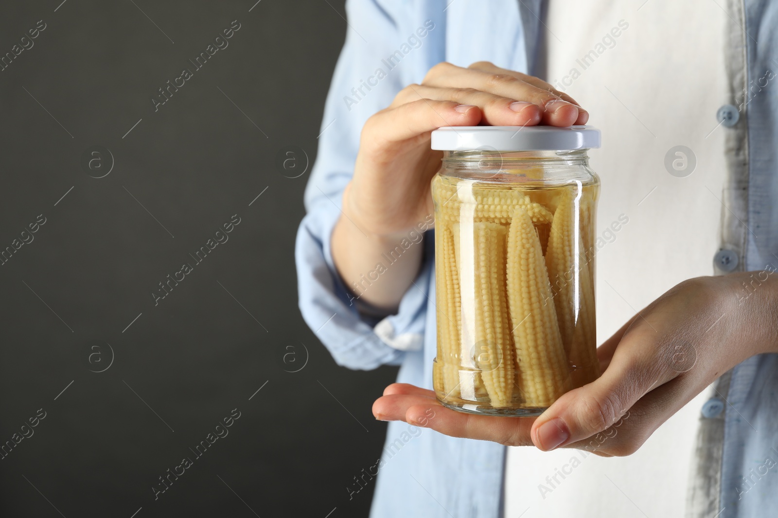 Photo of Woman with jar of tasty pickled yellow baby corns on gray background, closeup. Space for text