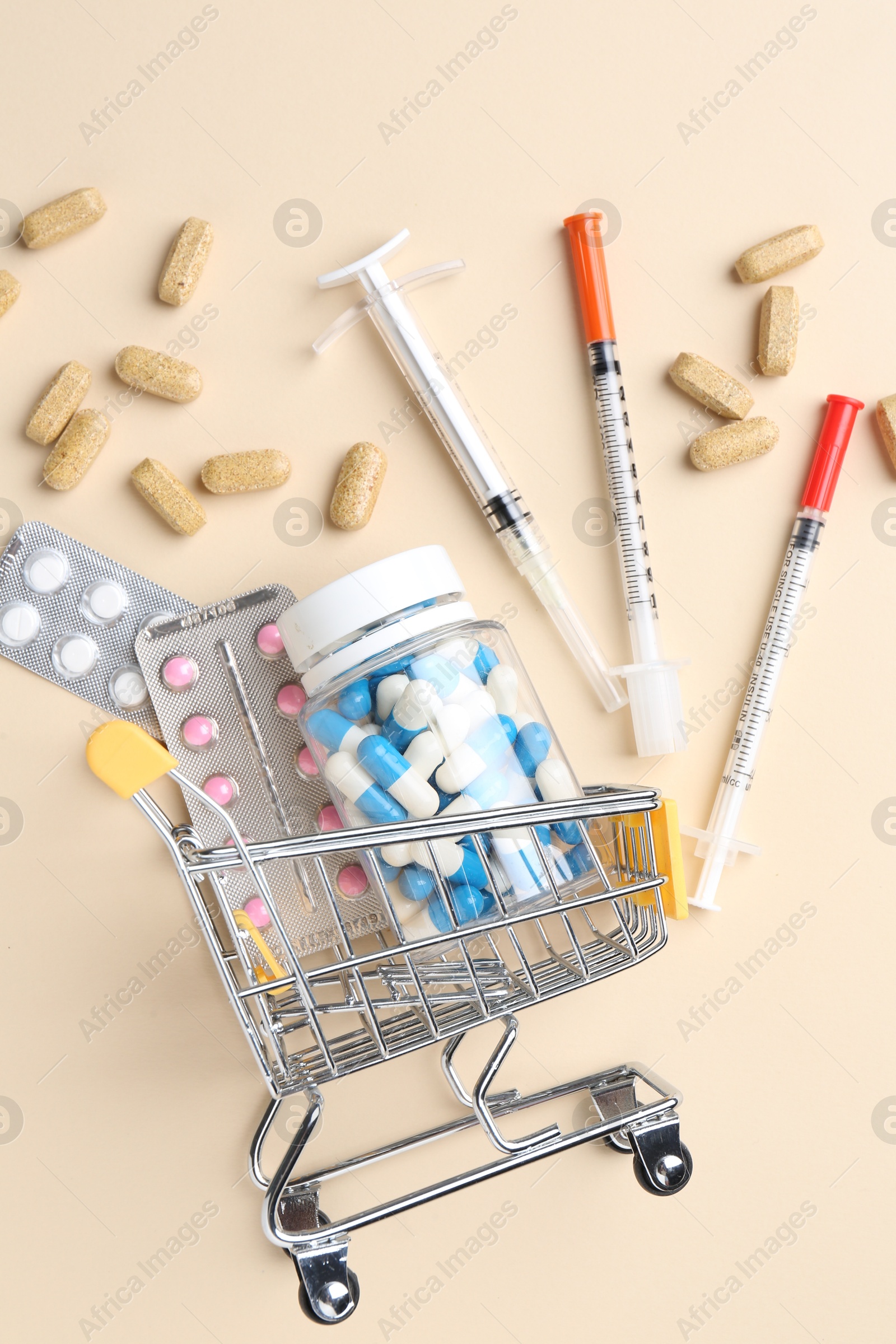 Photo of Pharmacist concept. Small shopping cart, syringes and pills on beige background, flat lay