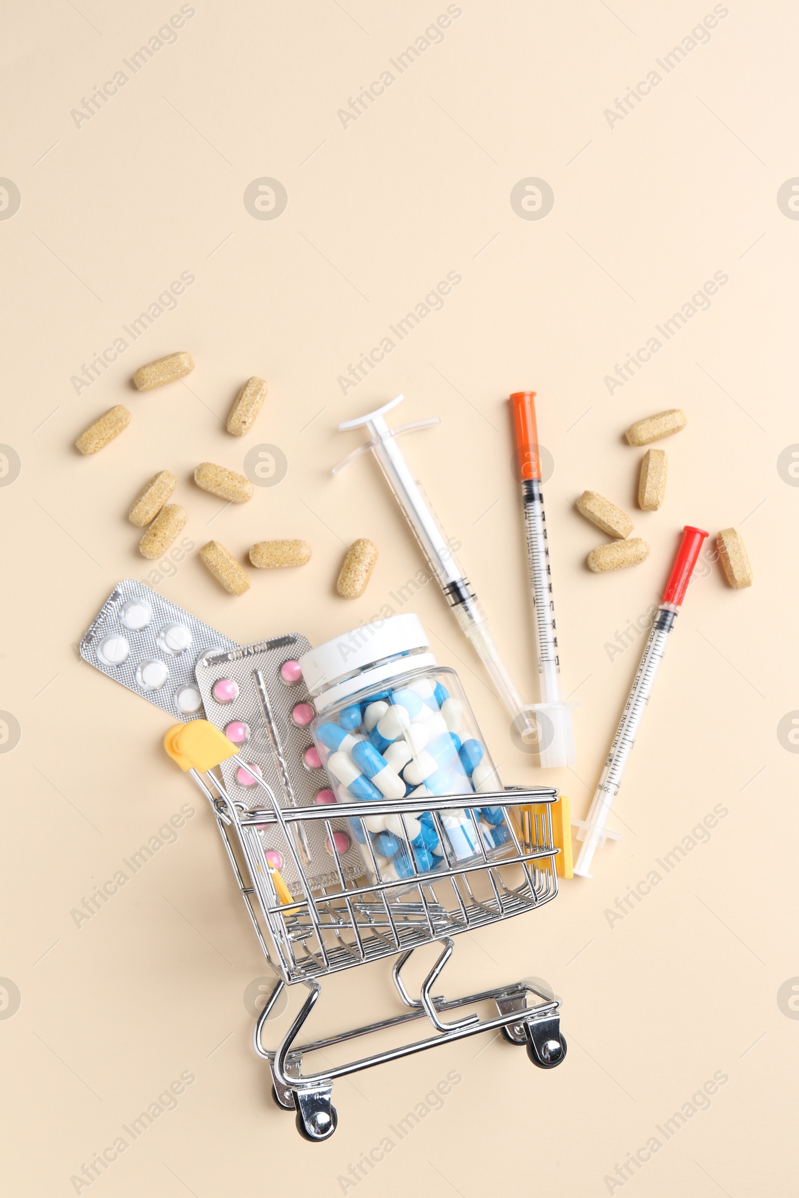 Photo of Pharmacist concept. Small shopping cart, syringes and pills on beige background, flat lay