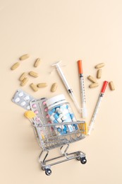 Pharmacist concept. Small shopping cart, syringes and pills on beige background, flat lay