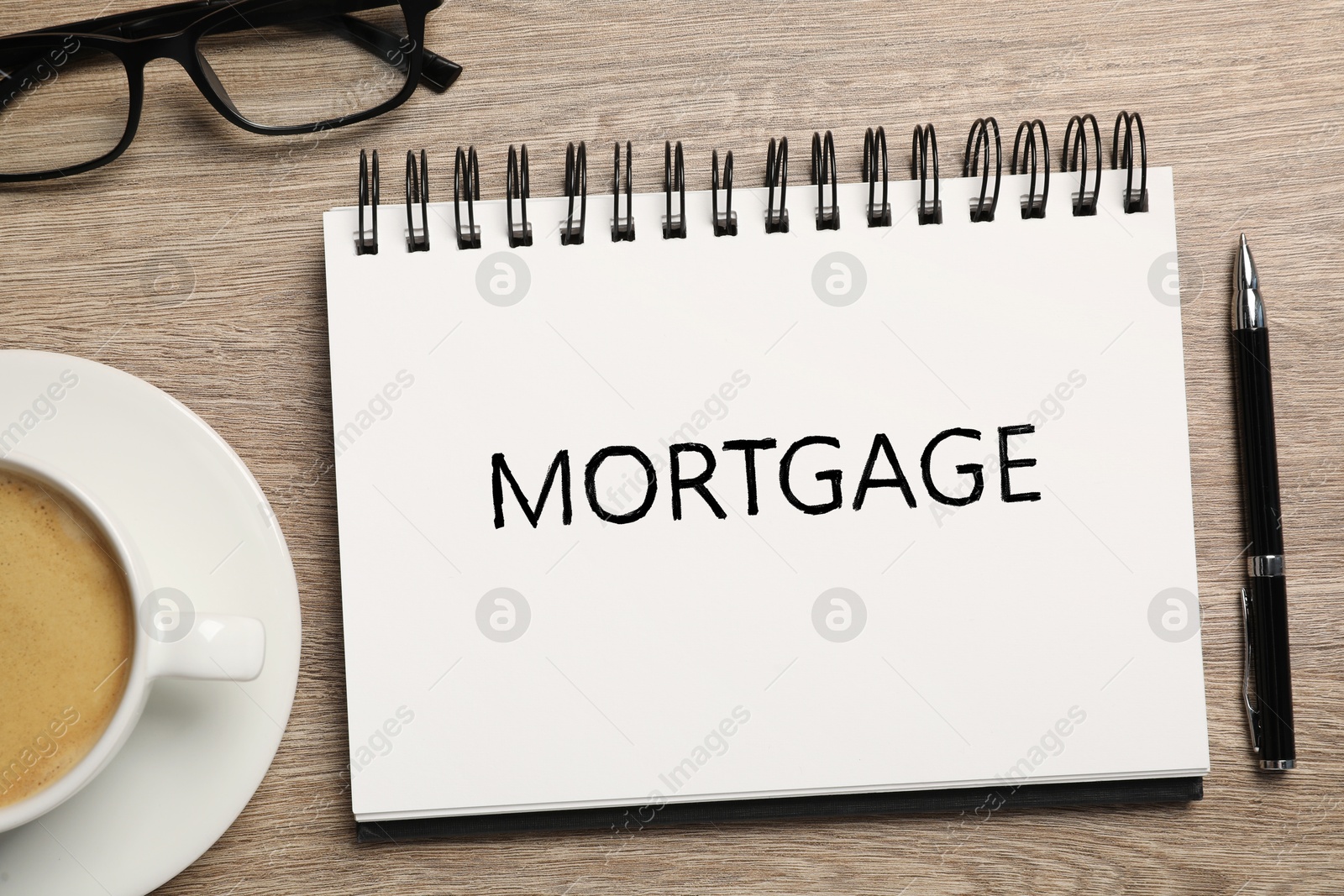 Image of Word Mortgage written in notebook, eyeglasses, pen and coffee on wooden table, flat lay