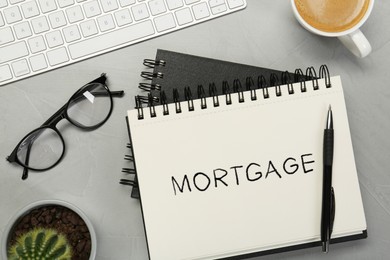 Image of Word Mortgage in notebook, eyeglasses and computer keyboard on grey table, flat lay