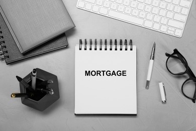 Image of Word Mortgage in notebook, eyeglasses and computer keyboard on grey table, flat lay