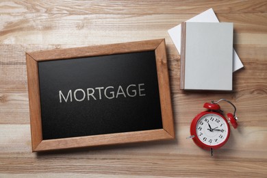 Image of Small chalkboard with word Mortgage, notebooks and alarm clock on wooden table, flat lay