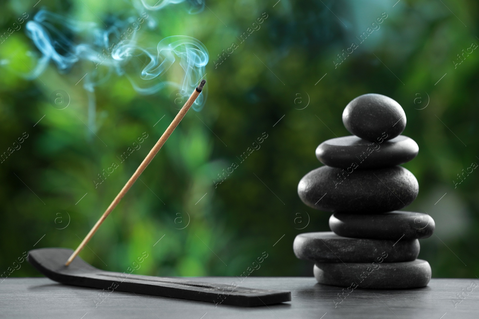 Photo of Incense stick smoldering in holder and spa stones on wooden table outdoors