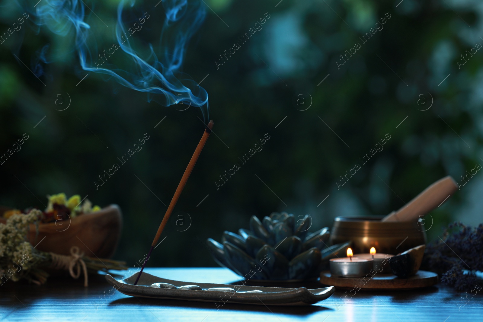 Photo of Incense stick smoldering in holder, burning candles, dry flowers and Tibetan singing bowl on wooden table outdoors