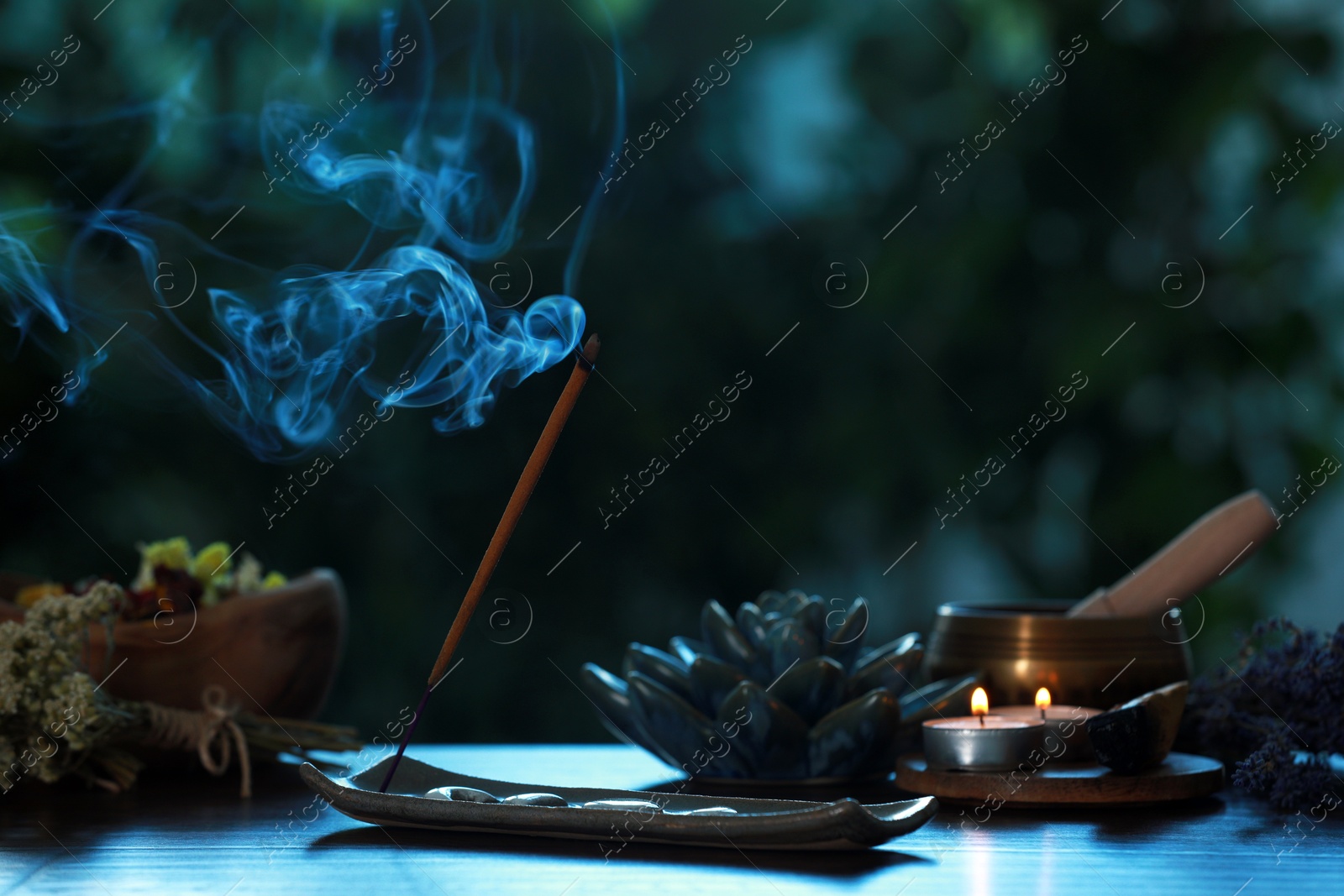 Photo of Incense stick smoldering in holder, burning candles, dry flowers and Tibetan singing bowl on wooden table outdoors