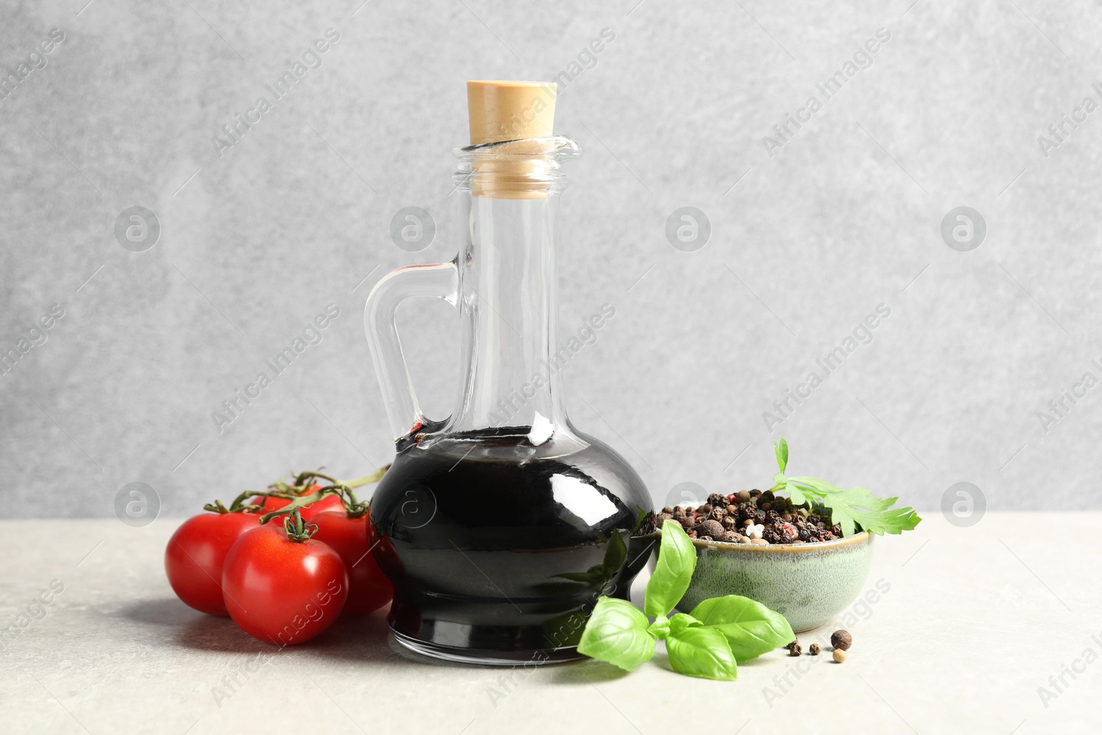 Photo of Balsamic vinegar in glass jug, tomatoes, herbs and spices on gray textured table