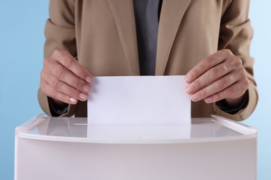 Woman putting her vote into ballot box against light blue background, closeup