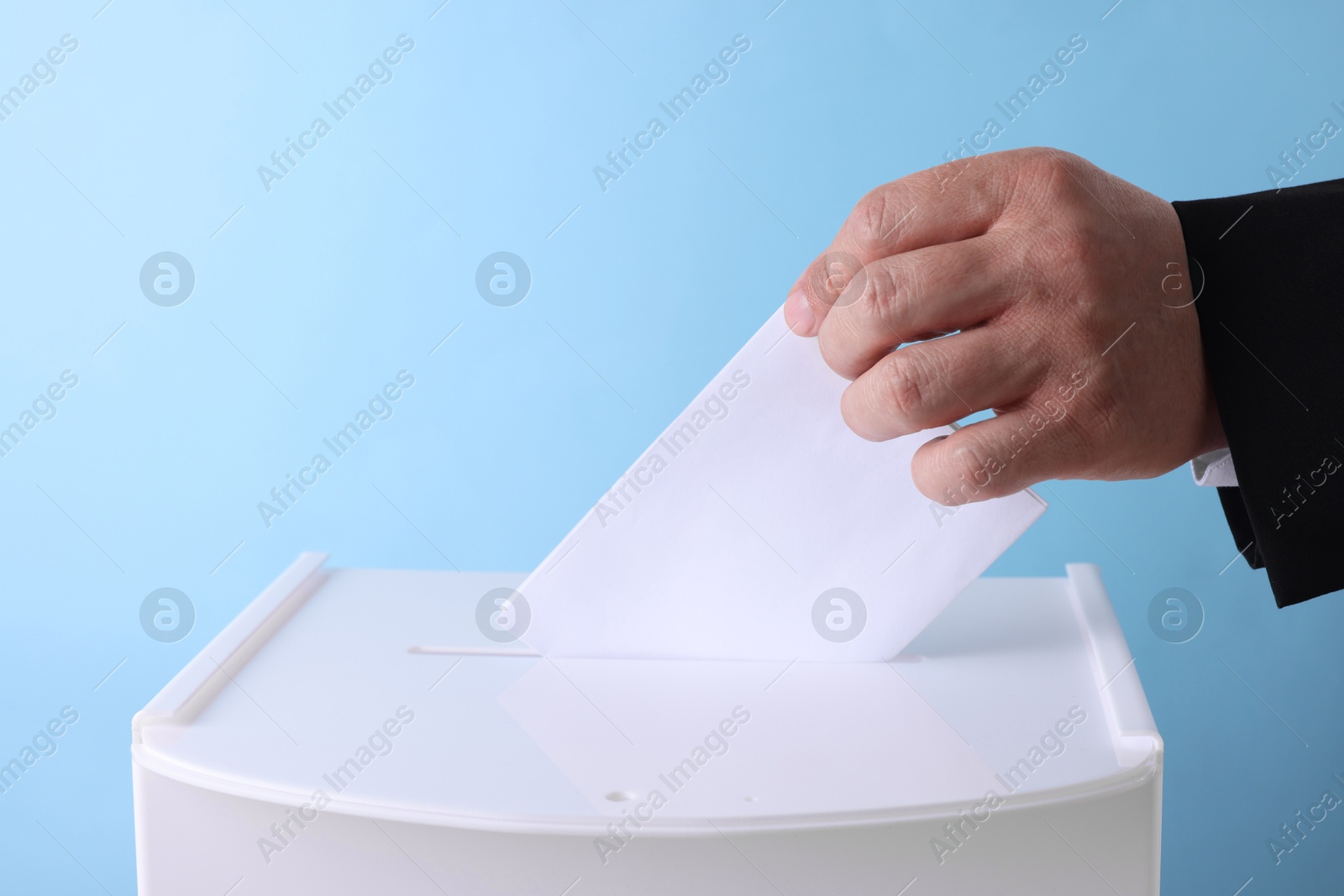 Photo of Man putting his vote into ballot box against light blue background, closeup