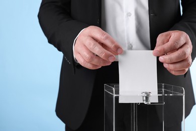 Man putting his vote into ballot box against light blue background, closeup